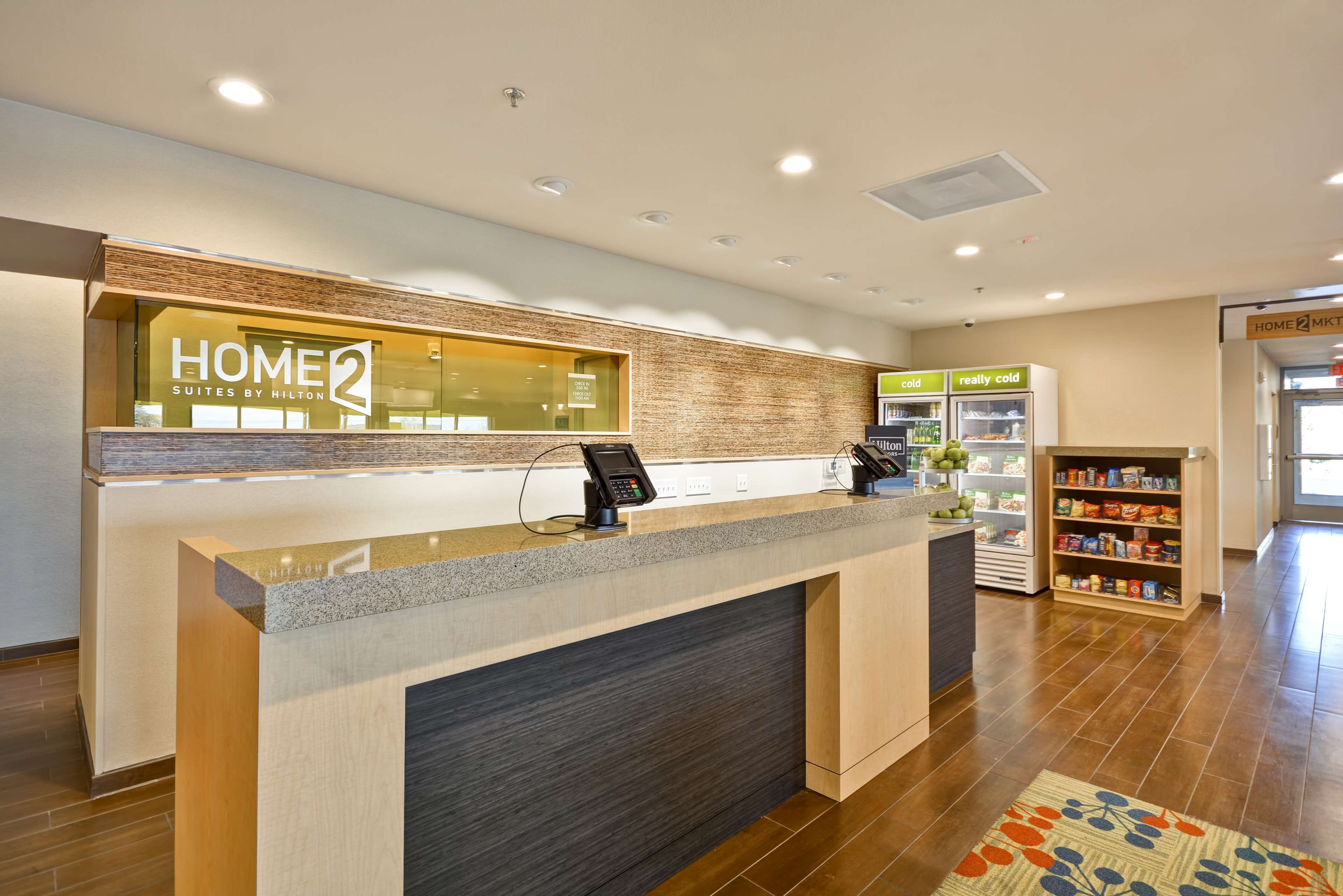 Un espace d’accueil d’hôtel comprenant un comptoir de réception, un écran numérique, un kiosque en libre-service et un réfrigérateur rempli de boissons.