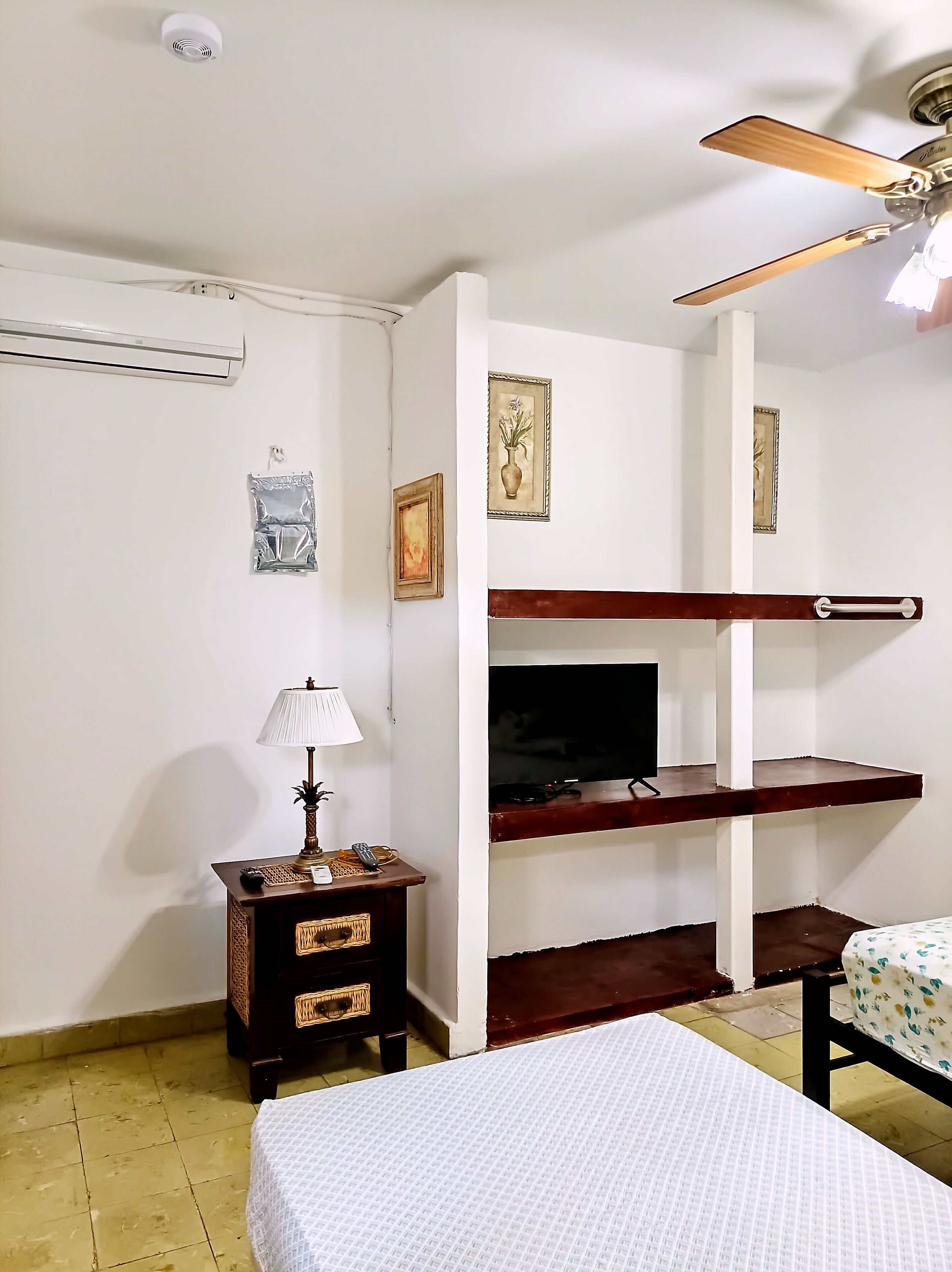 Una habitación con una cama, un televisor en un estante de madera, una mesita con una lámpara y un ventilador de techo.