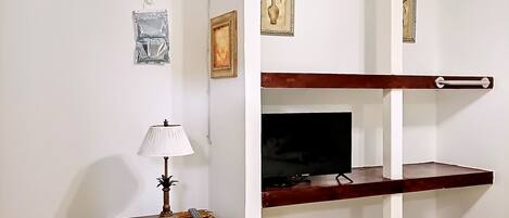 Una habitación con una cama, un televisor en un estante de madera, una mesita con una lámpara y un ventilador de techo.