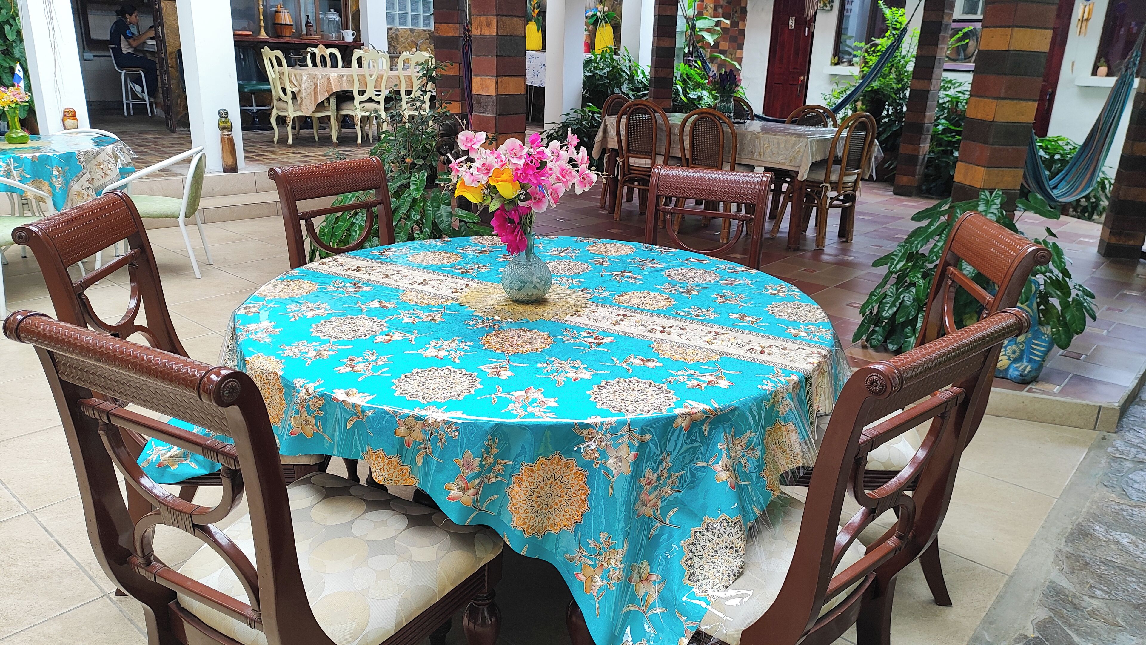 Une table ronde recouverte d’une nappe à motifs floraux, entourée de chaises en bois, dans une pièce dont le sol est carrelé et où l’on trouve des plantes en pot.