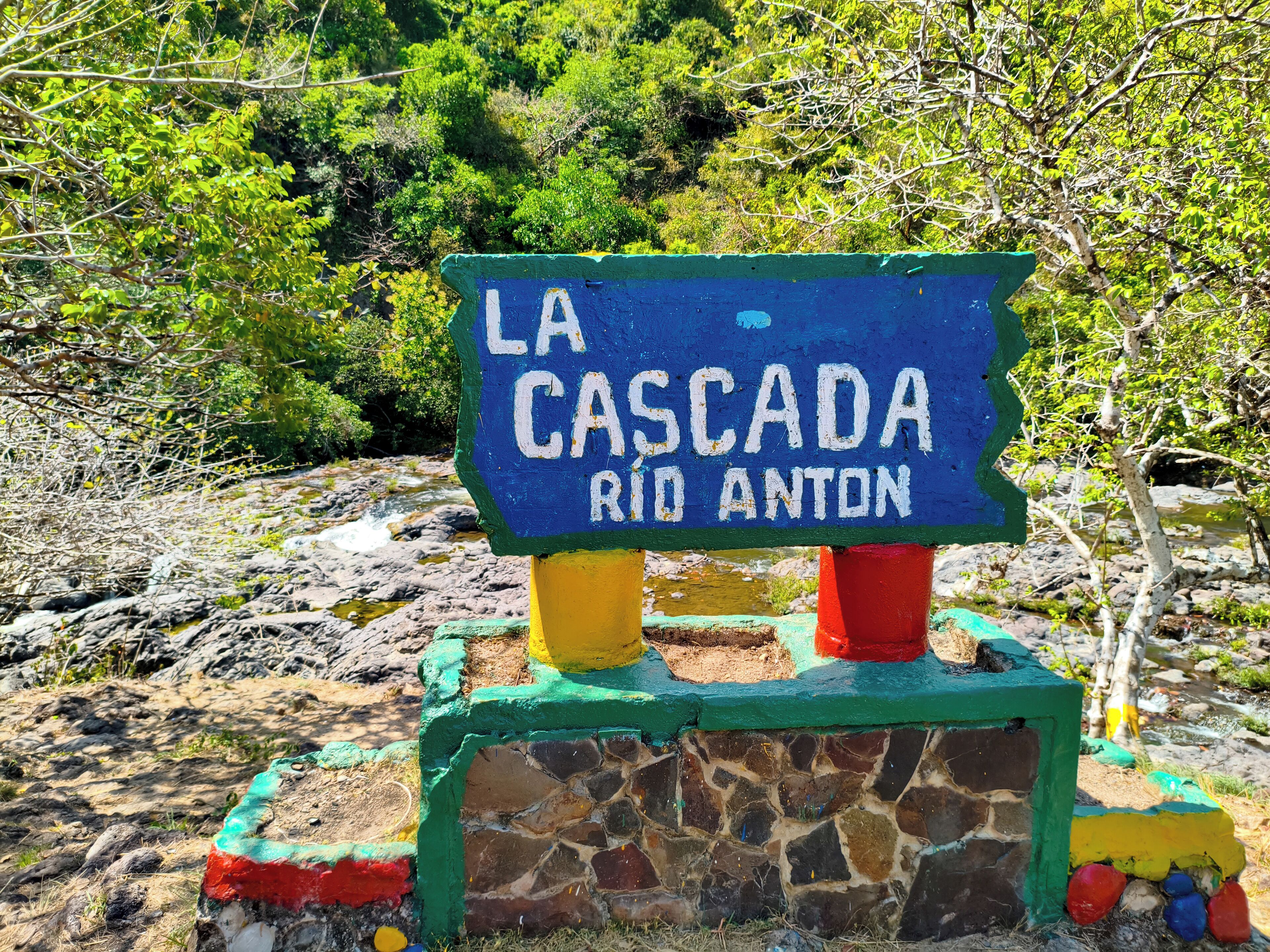 Un letrero que dice "LA CASCADA RIO ANTON", con base de piedra y soportes pintados de colores.