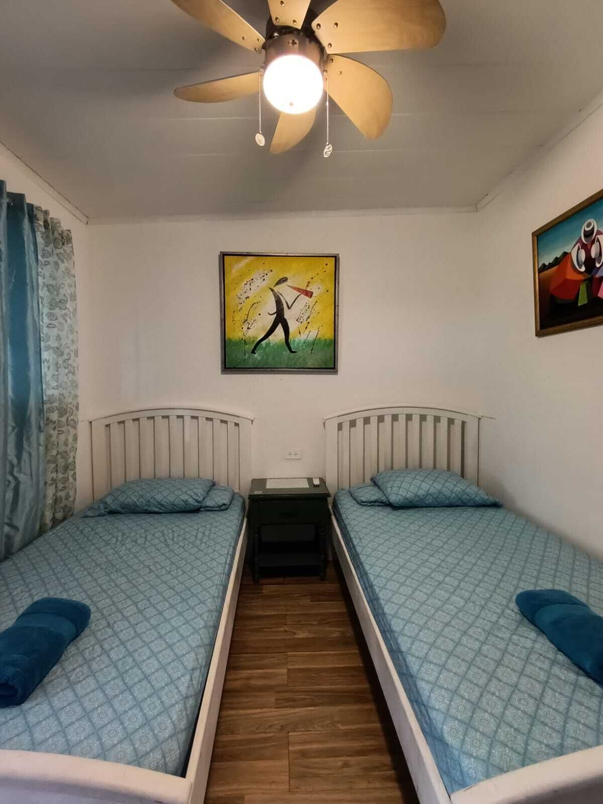 Een kamer met twee eenpersoonsbedden, een plafondventilator, een schilderij aan de muur en een raam met gordijnen.