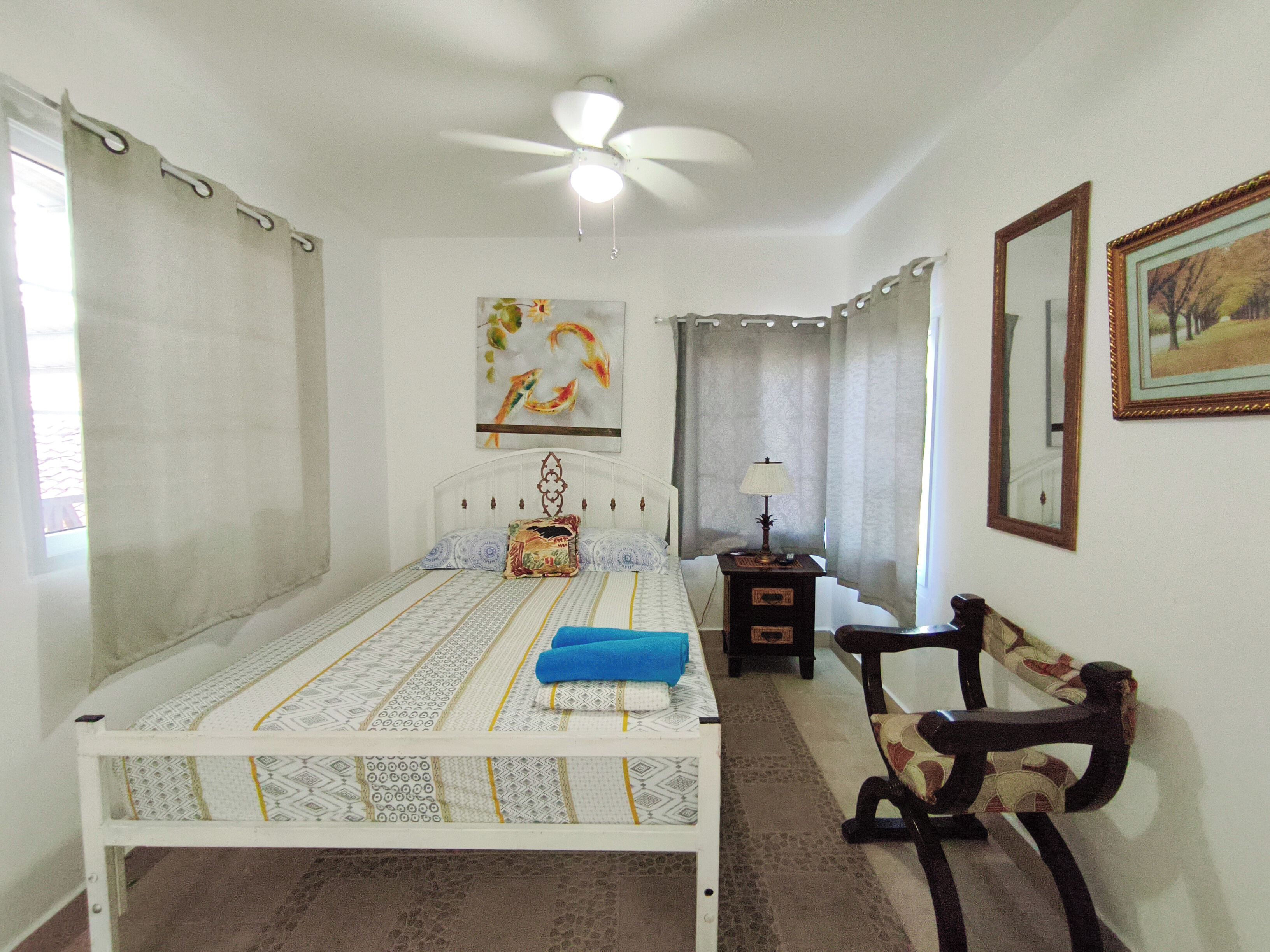 Un dormitorio con cama, una silla, una mesita de noche, ventilador de techo, un cuadro y un espejo.