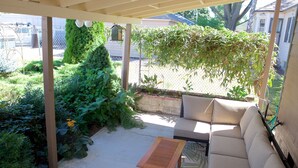Ein überdachter Patio-Bereich mit einer Couch und einem Holztisch, umgeben von üppiger Bepflanzung und Blick auf eine Wohngegend.