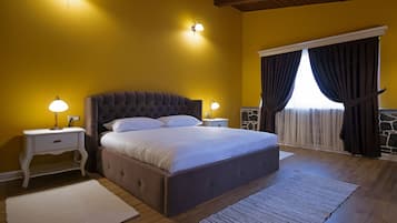 A bedroom with a large bed, two bedside lamps, a nightstand, a window with curtains, and a wooden floor.