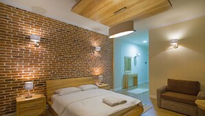 A bedroom with a brick wall, a wooden bed, a nightstand, a chair, and a bathroom visible through an open door. - Casa Gaçe Hotel (Qendër Bulgarec)