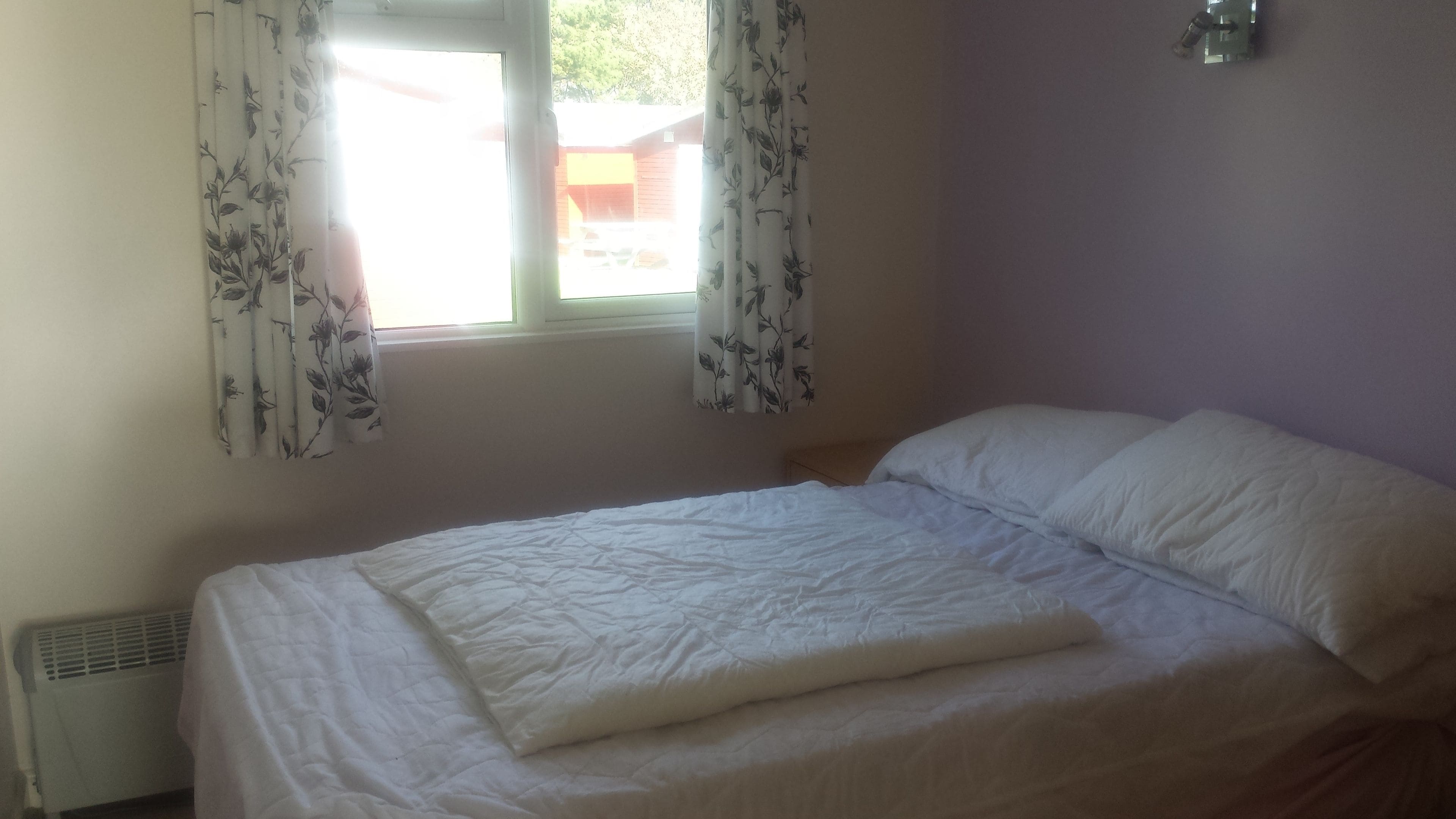 A bedroom with a bed, a window with floral curtains, and a wall-mounted light fixture.