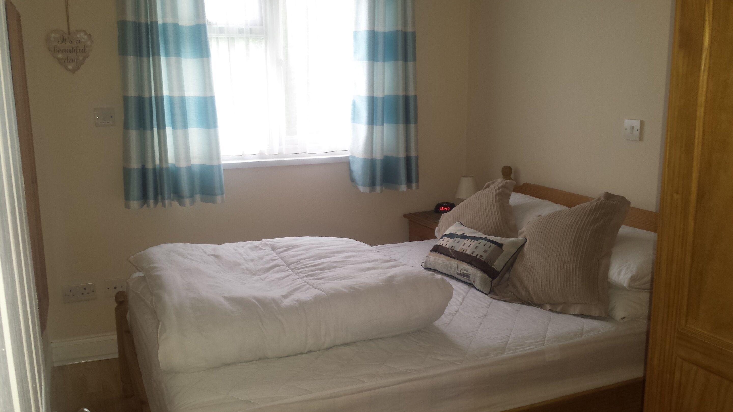 A neatly made bed with white bedding, a wooden headboard, and two pillows. There is a nightstand with a lamp and a clock. Blue and white striped curtains cover the window.