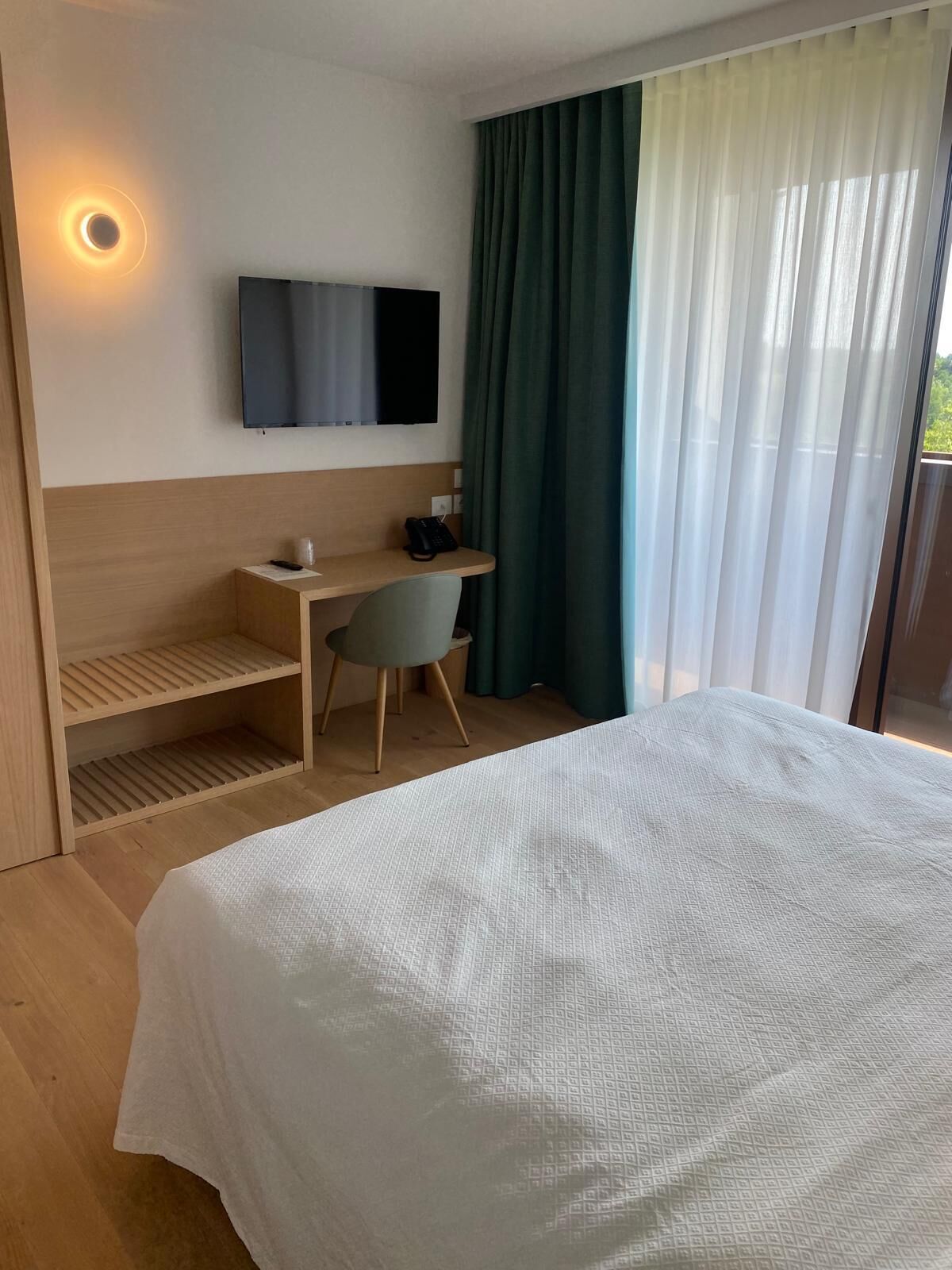 Quarto de hotel com cama, escrivaninha, cadeira, televisão presa à parede e janela com cortinas.