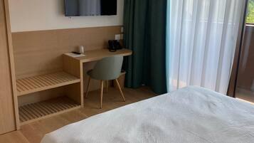 Quarto de hotel com cama, escrivaninha, cadeira, televisão presa à parede e janela com cortinas.