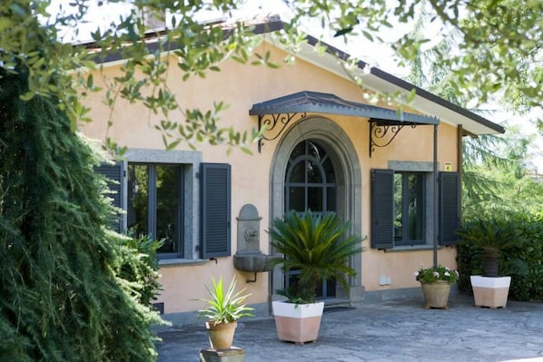 A building with a beige facade, black shutters, and a central arched doorway. There are potted plants in front of the building.