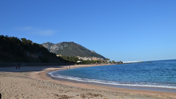 Una playa con una montaña al fondo, cielos despejados y algunas personas caminando.