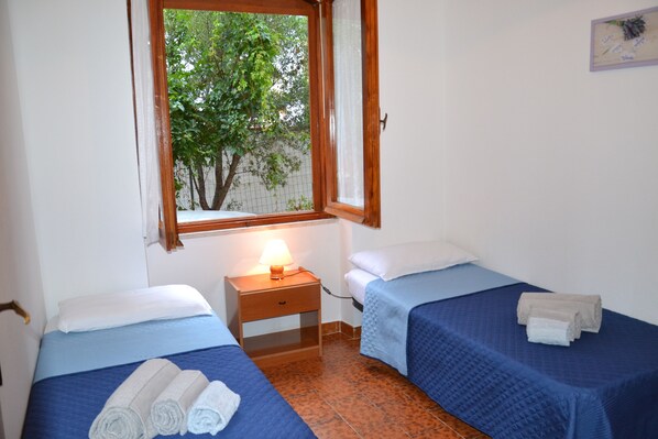 A room with two beds, a window with a view of trees, a nightstand with a lamp, and a wall-mounted picture.