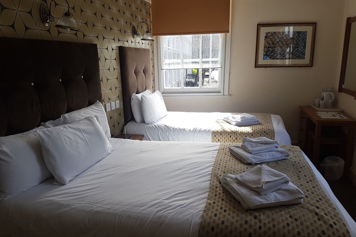 A hotel room with two beds, a window with a view, a small table with a kettle, and a wall-mounted lamp.