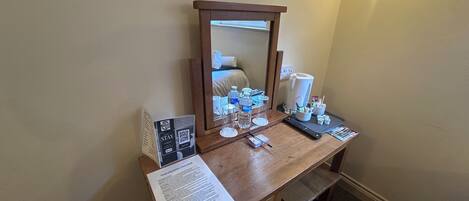 Een hotelkamer met een houten bureau, een spiegel, een aan de muur bevestigde televisie en een nachtkastje met toiletartikelen.