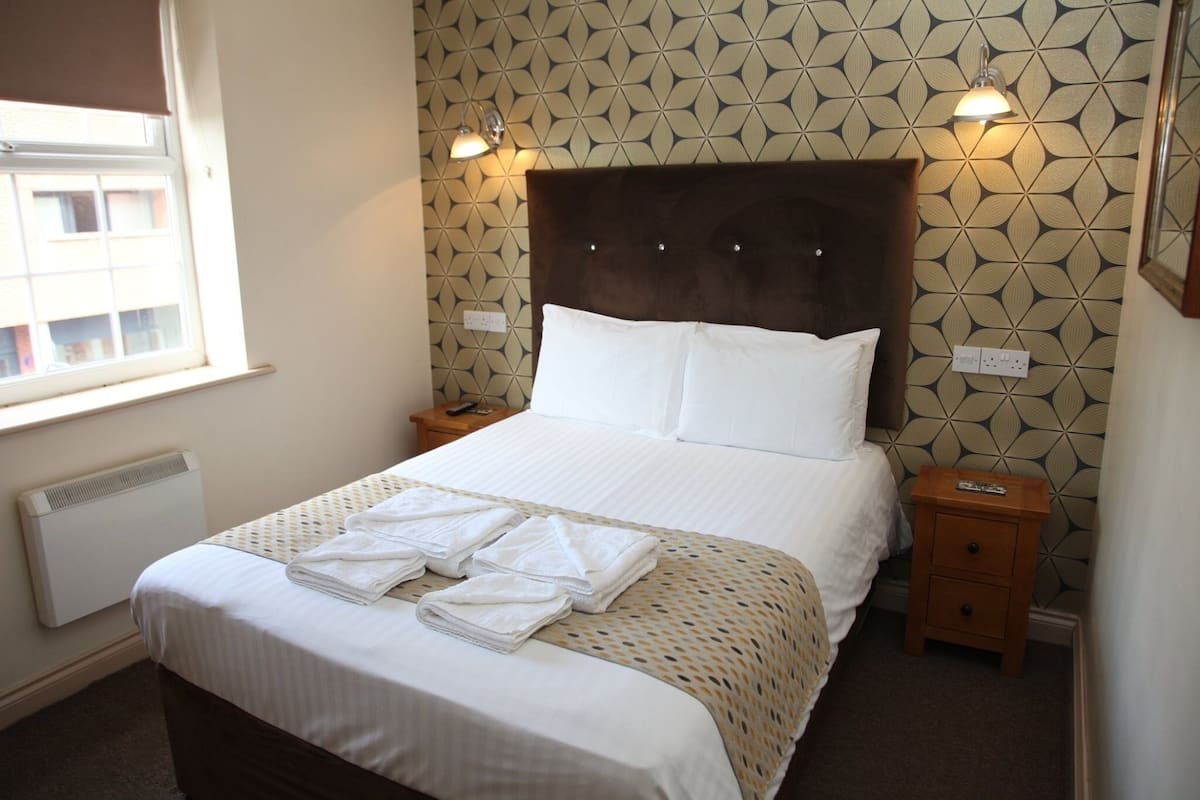 A neatly made bed with white linens and folded towels, a wooden nightstand with a lamp, and patterned wallpaper.
