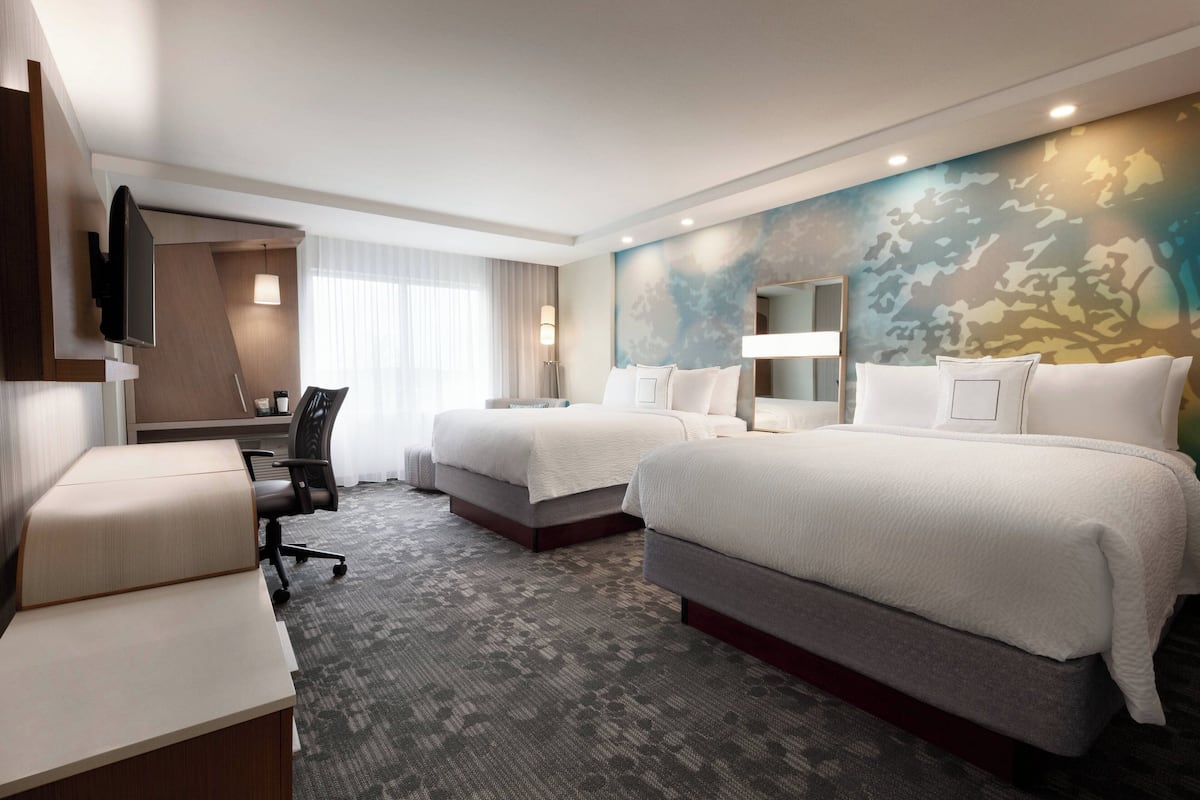 Ein Hotelzimmer mit zwei Betten, einem Schreibtisch, einem Stuhl, einem Fernseher und einem großen Fenster mit Vorhängen.