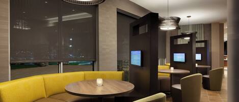 Un moderno vestíbulo de hotel con zona de estar que incluye un sofá amarillo y mesas redondas, ventanas amplias con vista a la ciudad e iluminación contemporánea.
