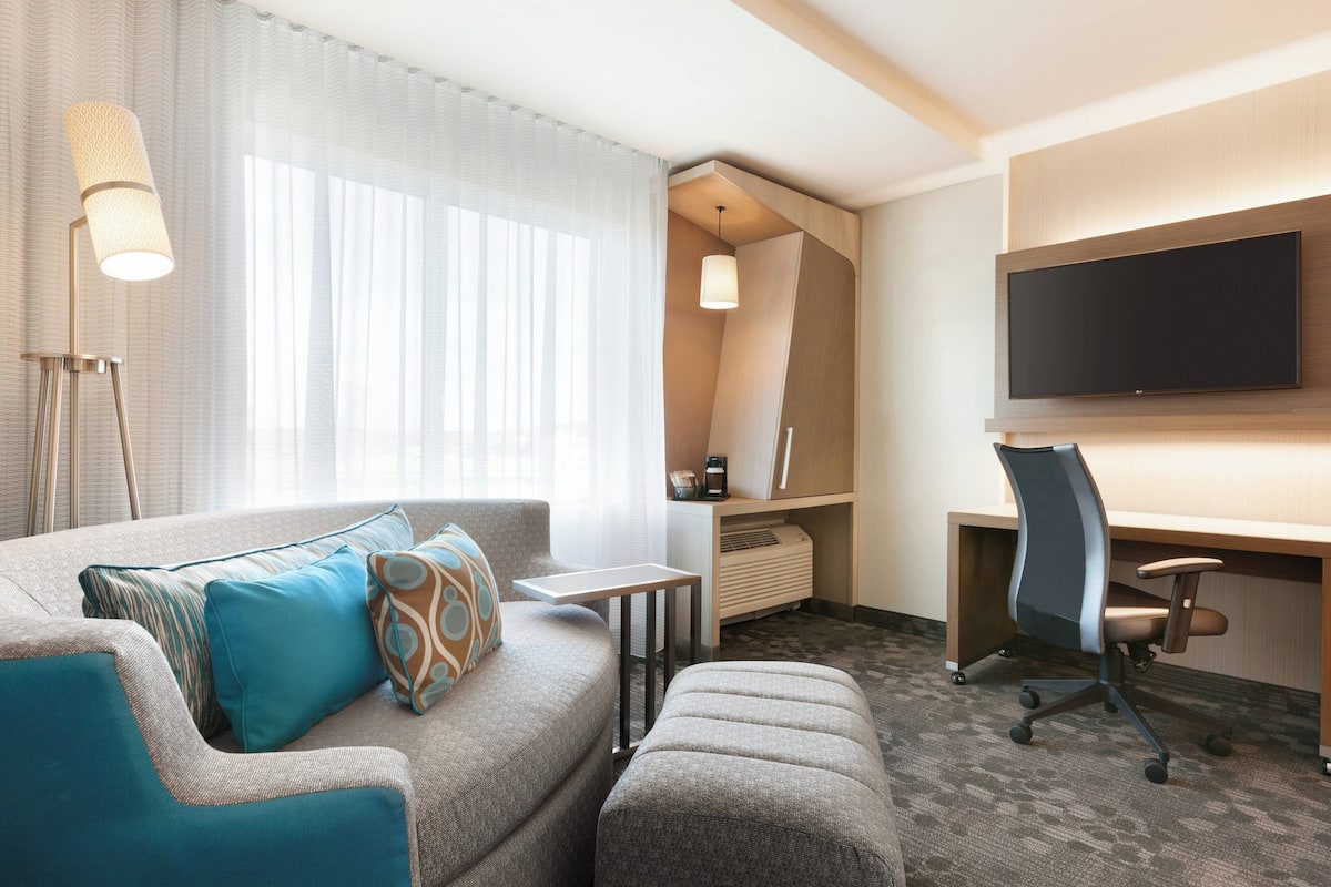 Ein modernes Hotelzimmer mit Sofa, Hocker, Schreibtisch und Flachbildfernseher.