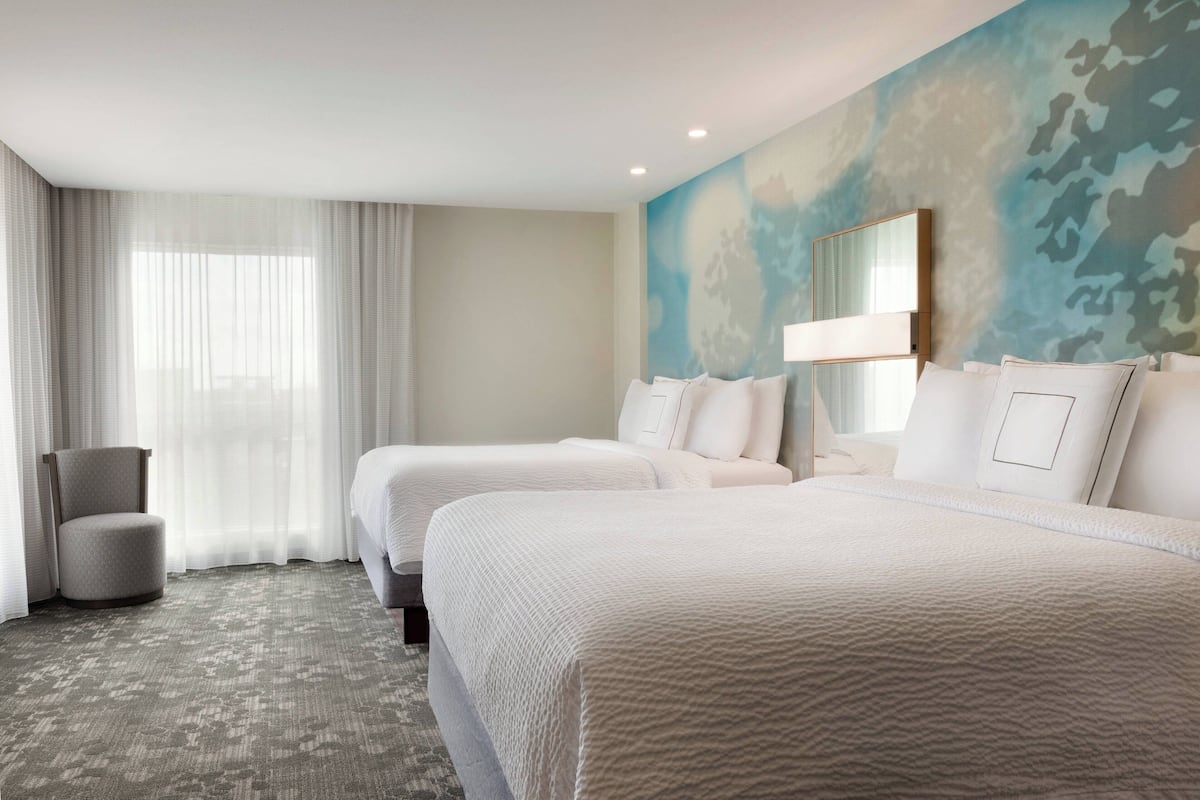 Ein Hotelzimmer mit zwei Betten, einem Sessel und einem großen Fenster mit durchsichtigen Vorhängen.
