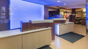 Un área de recepción de hotel con un diseño moderno, que incluye un mostrador de recepción, un amplio ventanal y una zona de estar con mesas y sillas.