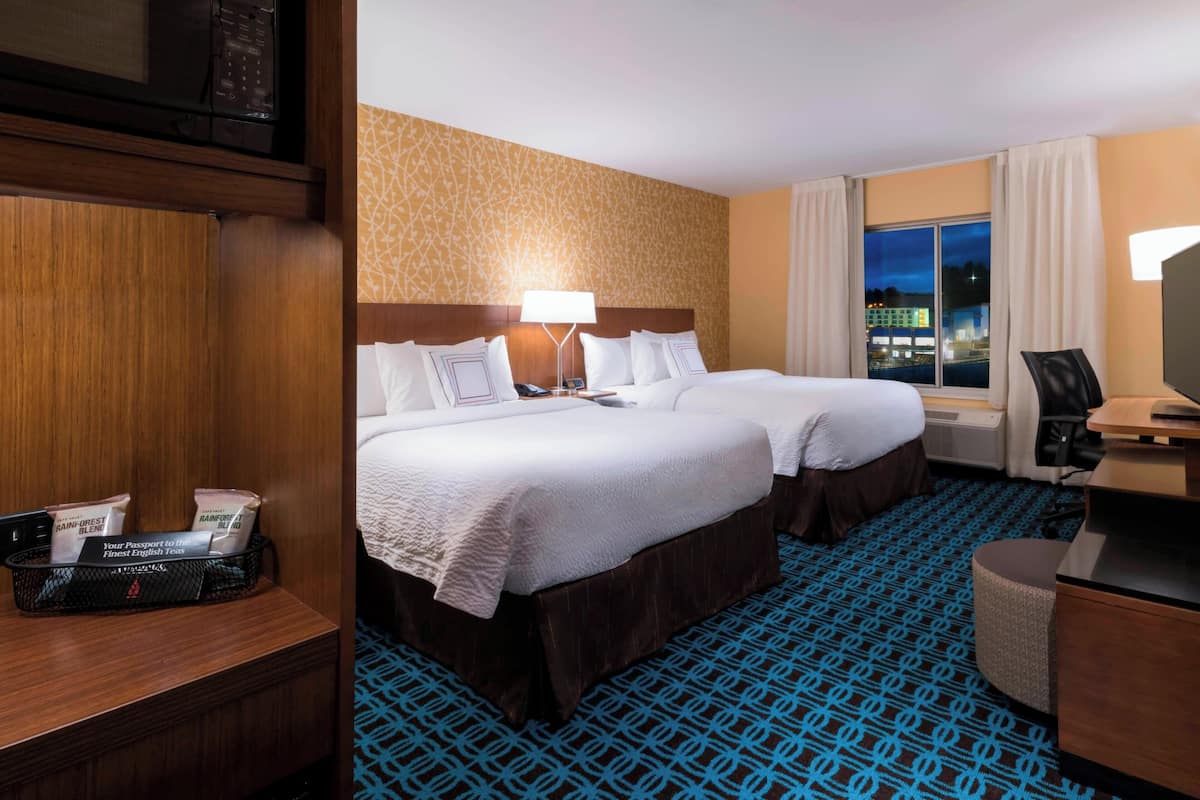 A hotel room with two beds, a desk with a chair, a window with a view of the city, and a minibar.