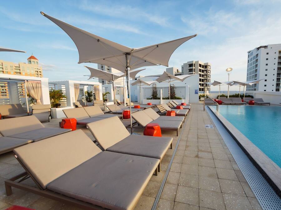 A rooftop pool area with sun loungers and umbrellas, a clear blue sky, and modern buildings in the background.