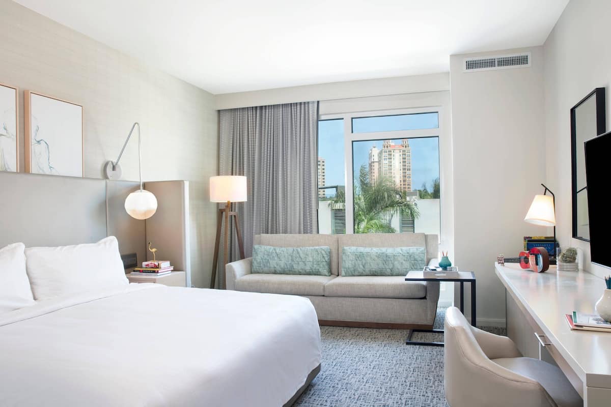 Hotelkamer met een groot bed, een bank, een bureau en uitzicht op palmbomen en gebouwen.