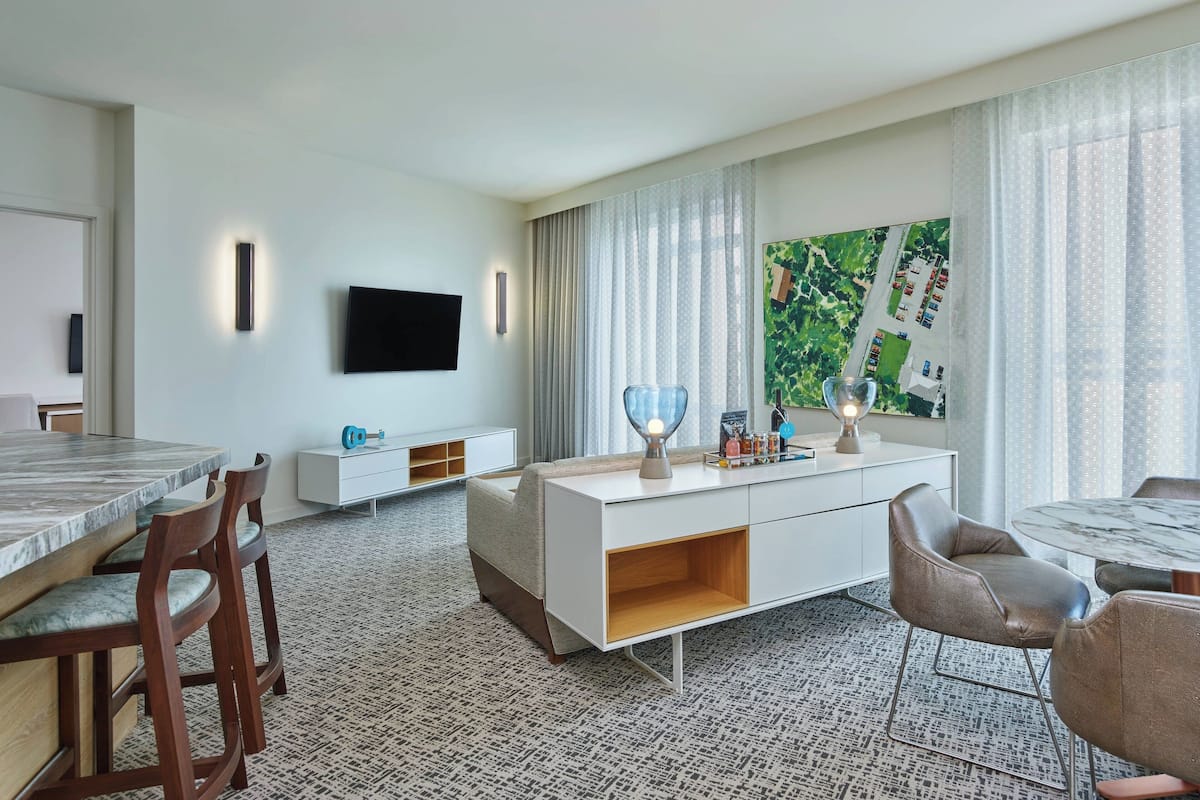 Een moderne hotelkamer met een zithoek, een flatscreen televisie aan de muur, een groot raam met uitzicht op het landschap, en een witte console met een lamp en decoratieve objecten.