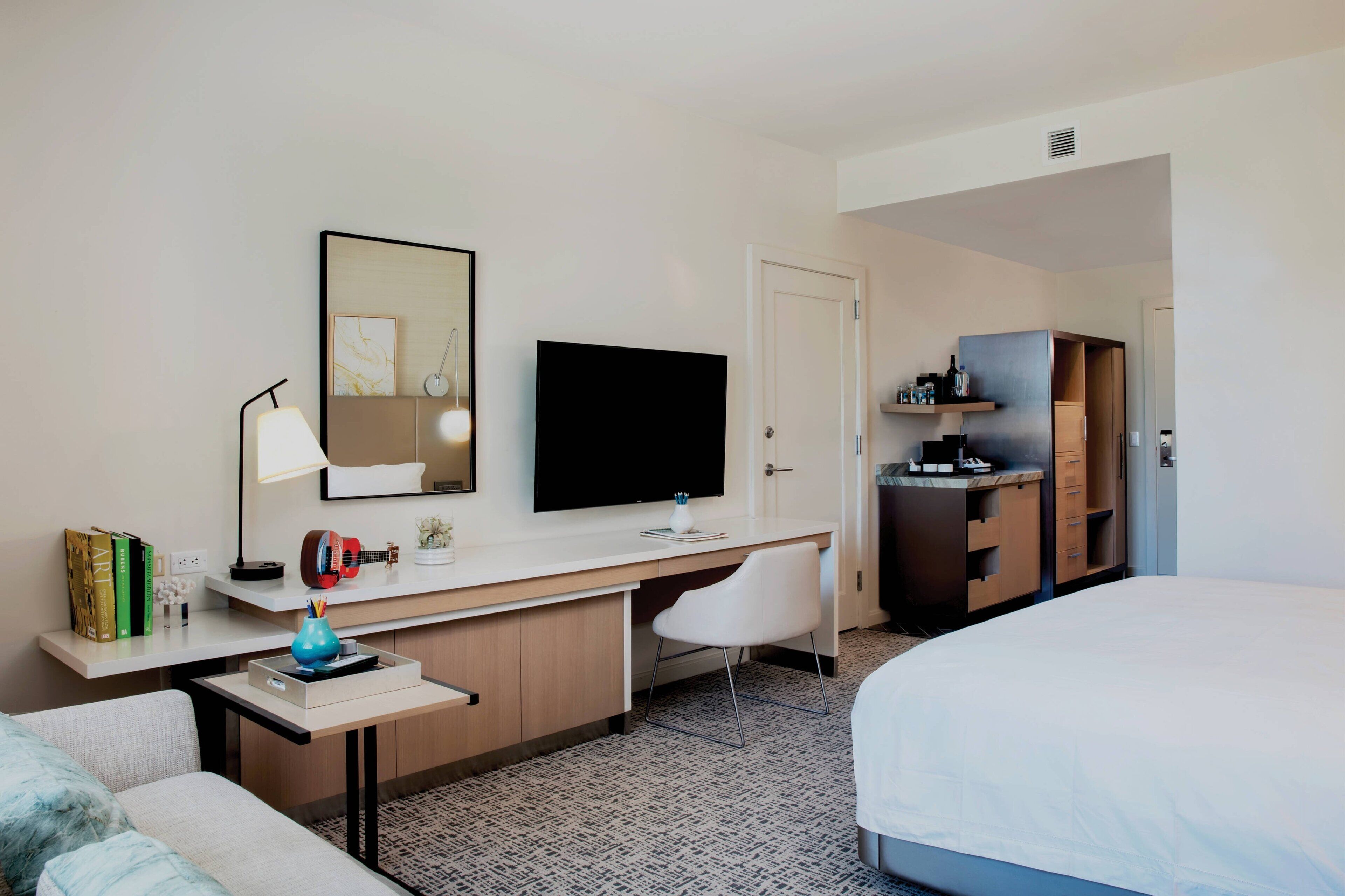A hotel room with a bed, a desk with a flat-screen TV, a chair, a small table, and a wardrobe.