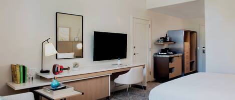 A hotel room with a bed, a desk with a flat-screen TV, a chair, a small table, and a wardrobe.