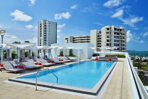 Ein Pool auf dem Dach mit Liegestühlen und Sonnenschirmen, umgeben von modernen Gebäuden und blauem Himmel.