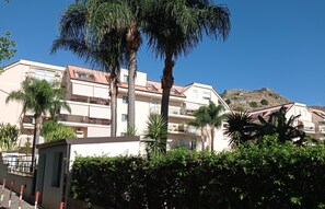 Um edifício de vários andares com varandas, rodeado por vegetação exuberante e uma colina ao fundo.
