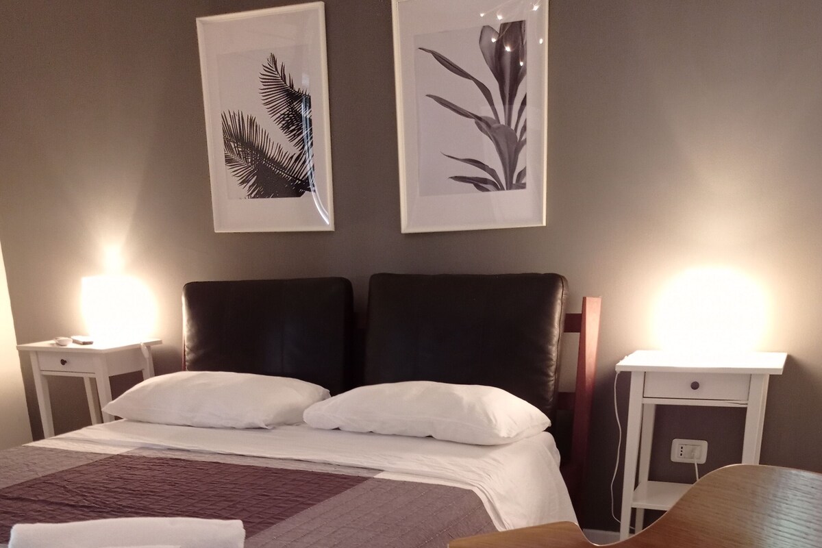 Quarto de hotel bem organizado, com uma cama, duas mesinhas de cabeceira, uma cadeira e duas fotos emolduradas na parede.