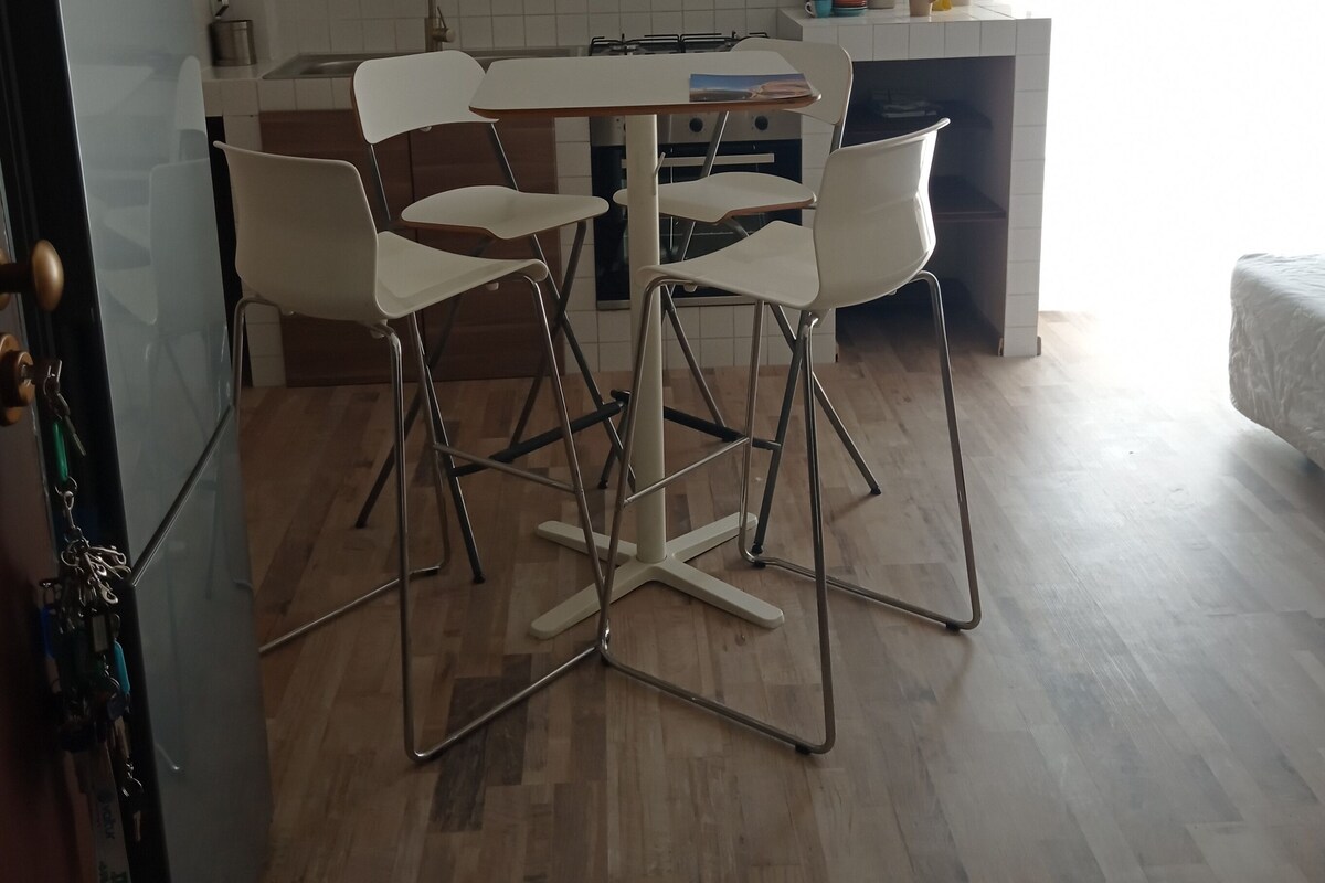 Uma cozinha com uma mesa de jantar, quatro cadeiras, geladeira, armários e uma janela com cortinas.
