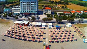 A beachside hotel with multiple beach umbrellas and sun loungers, a parking area, and a building with a red roof in the background. - Hotel Atlantic (Senigallia)