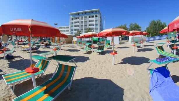 A beach with multiple sun loungers and umbrellas, a building in the background, and clear skies.