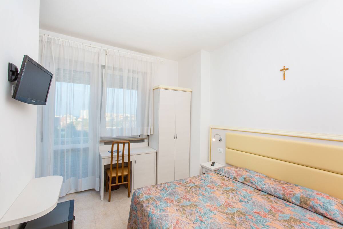A bedroom with a bed, a desk, a chair, a wardrobe, a television, and a cross on the wall.