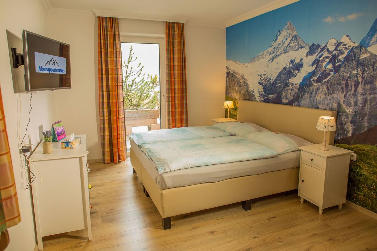 A bedroom with a bed, a nightstand, a TV on the wall, and a mountain painting.