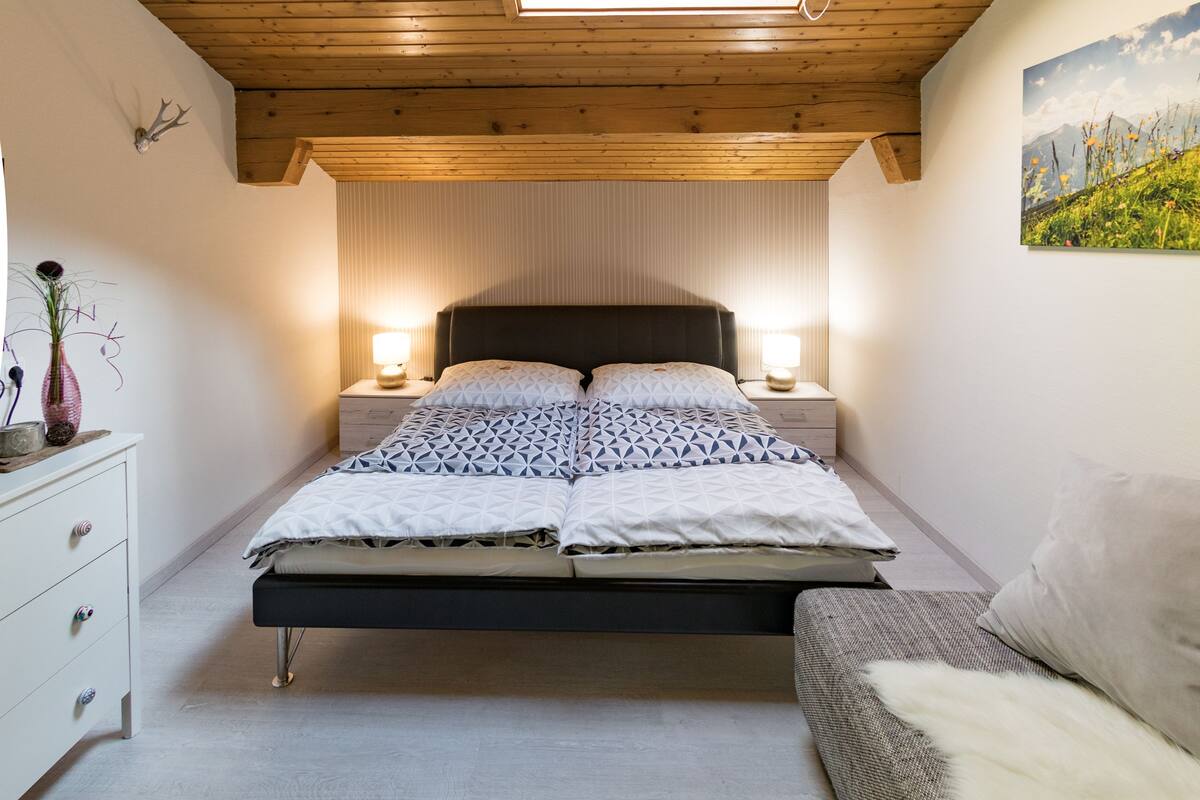 A bedroom with a sloped ceiling, a skylight, a bed with patterned bedding, bedside lamps, a dresser, and a wall-mounted deer head decoration.