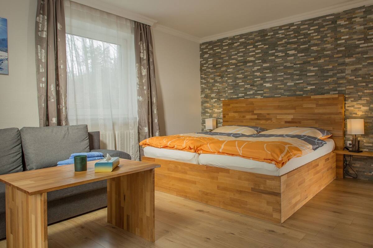 A bedroom with a wooden bed, a wooden nightstand, a bedside table, a grey sofa, and a window with curtains.