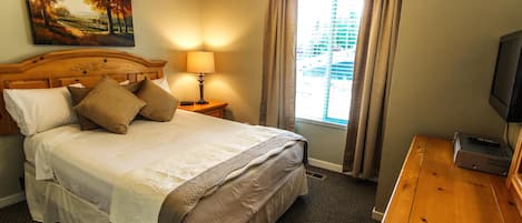 Habitación de hotel con cama, mesita de noche con lámpara, televisor y ventana con cortinas.