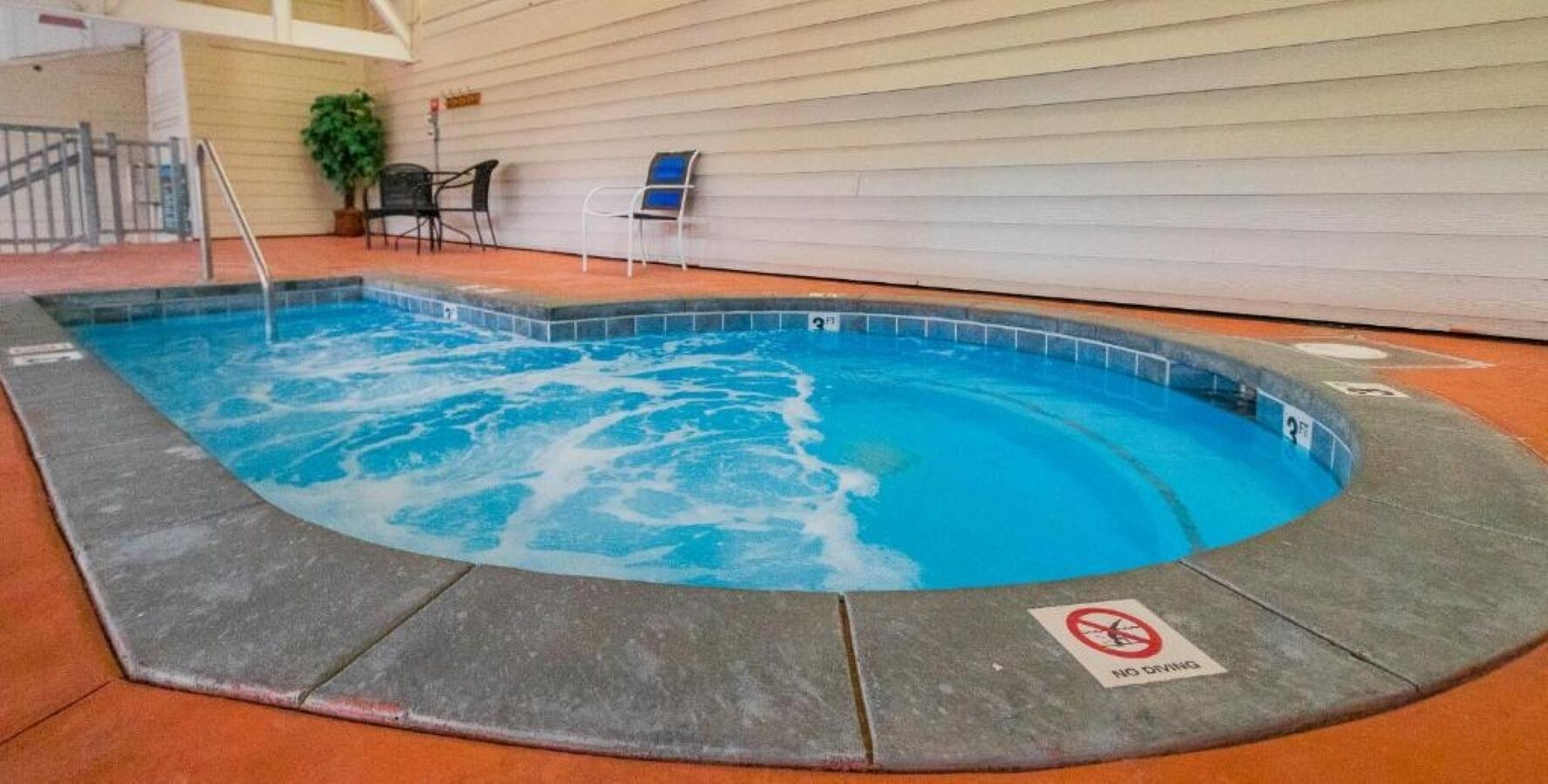 An indoor swimming pool with a 'No Diving' sign, surrounded by a concrete deck and a covered area with chairs.