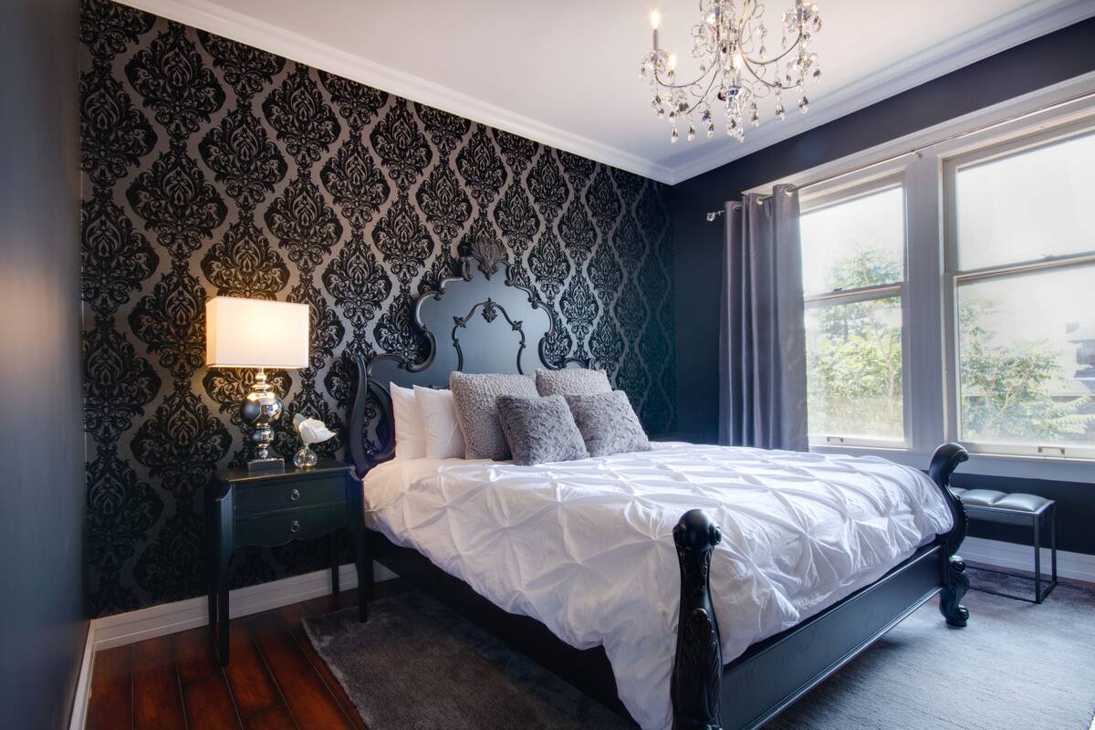 A bedroom with a dark patterned wallpaper, a white bed with multiple pillows, a bedside lamp, and a window with curtains.