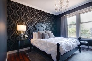 A bedroom with a dark patterned wallpaper, a white bed with multiple pillows, a bedside lamp, and a window with curtains. - Ember Hostel (Denver)