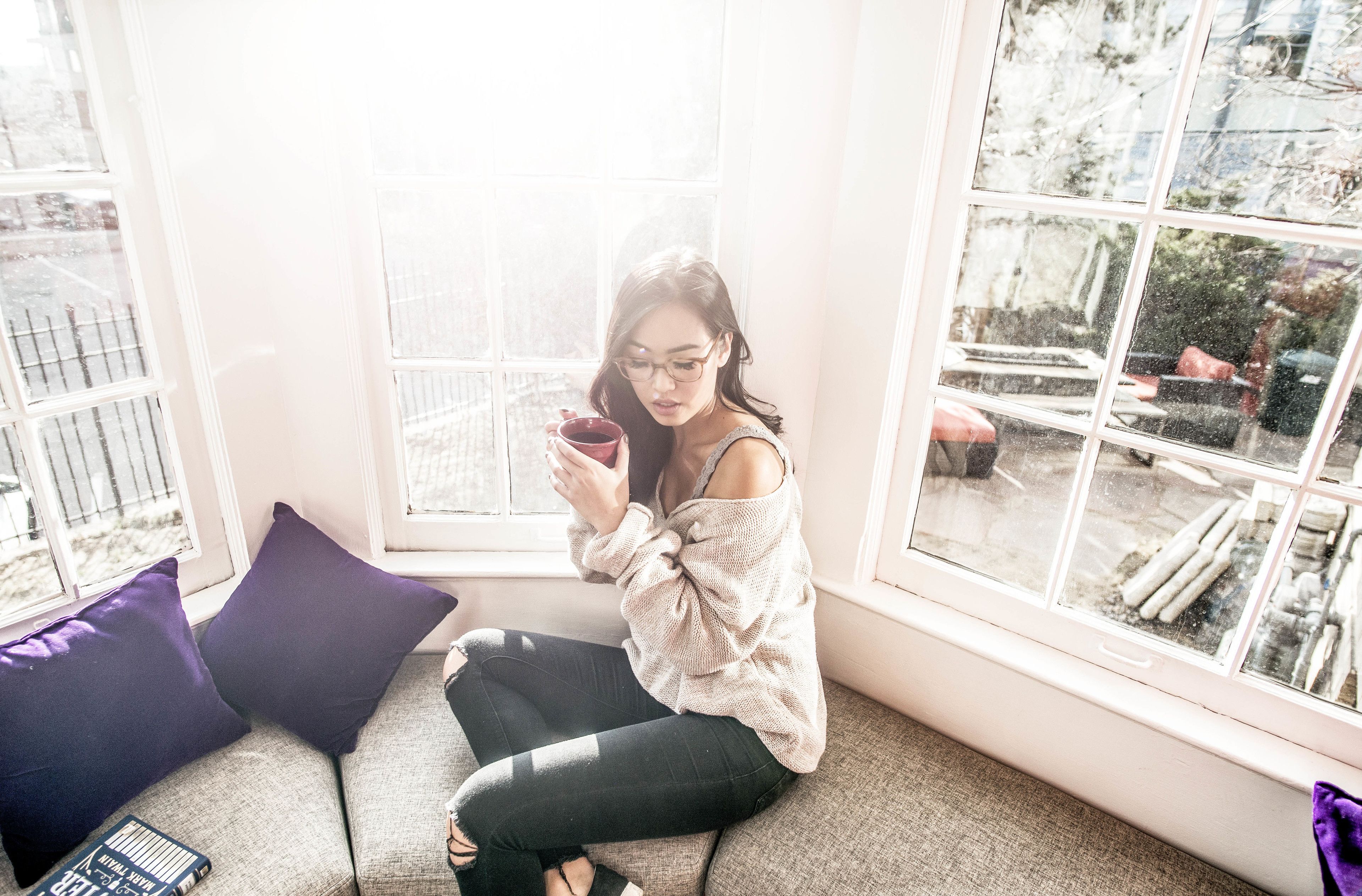 A person sitting on a grey sofa with a window behind them, holding a cup.