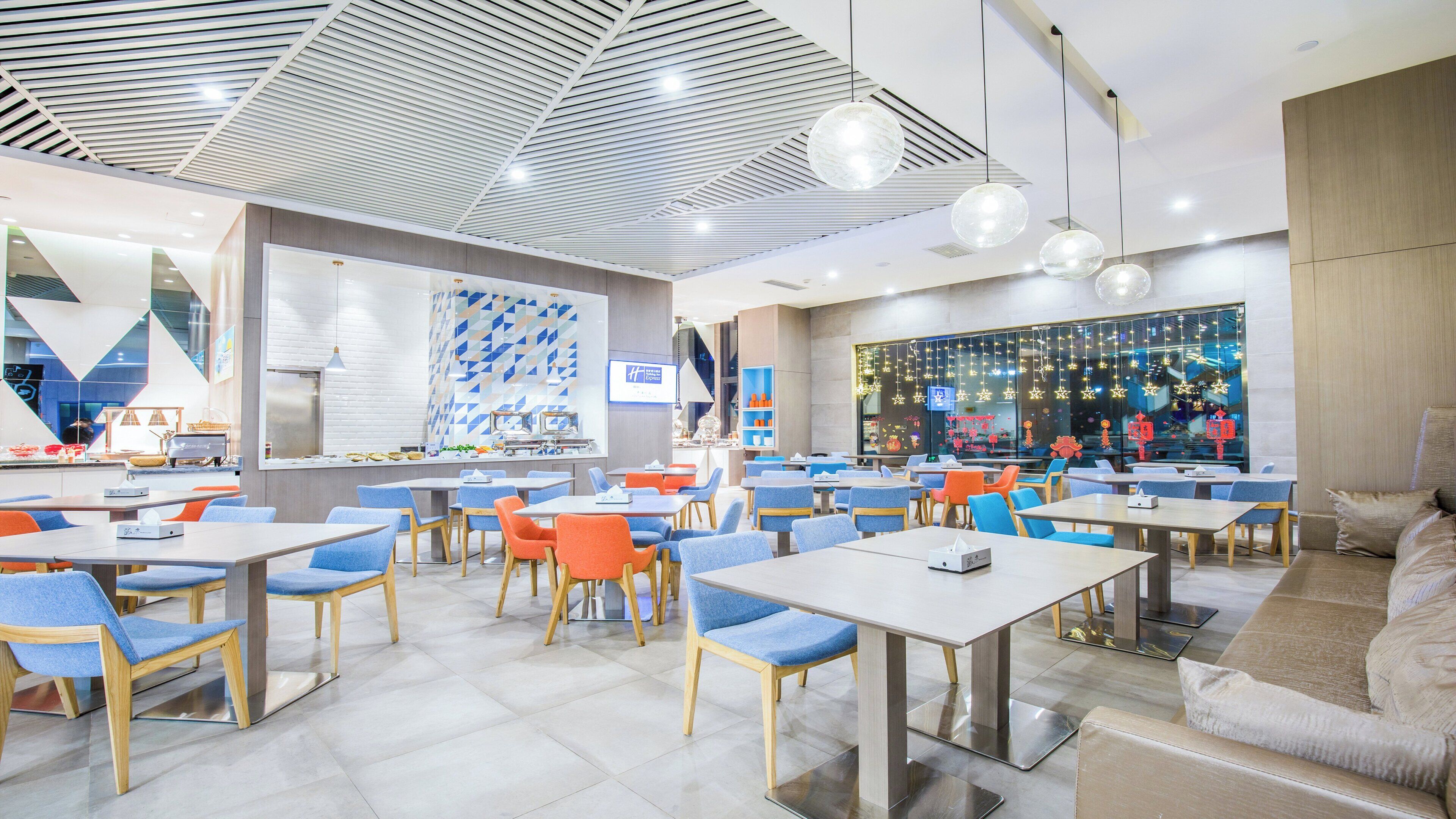 Un interior de restaurante moderno con varias mesas y sillas, una zona de cocina y un ventanal con adornos festivos.
