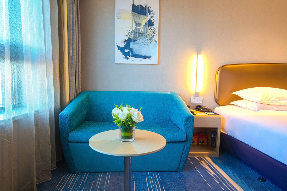 Ein Hotelzimmer mit einem blauen Sofa, einem kleinen runden Tisch mit Blumenvase, einem Bett mit weißer Bettwäsche, einem Nachttisch mit Lampe und einem Gemälde an der Wand.