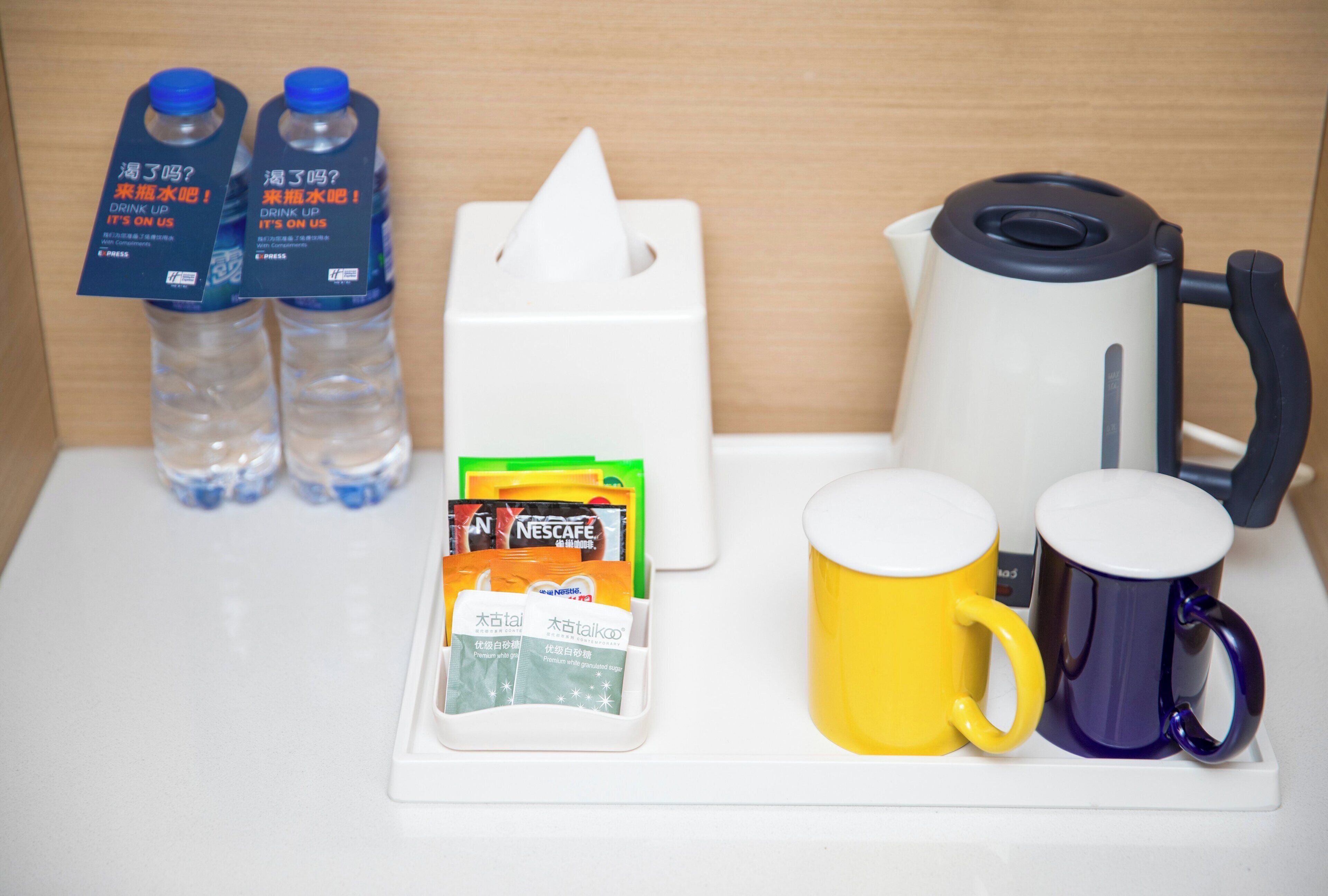 A hotel room's breakfast setup with a coffee maker, two mugs, a tissue box, and bottled water.