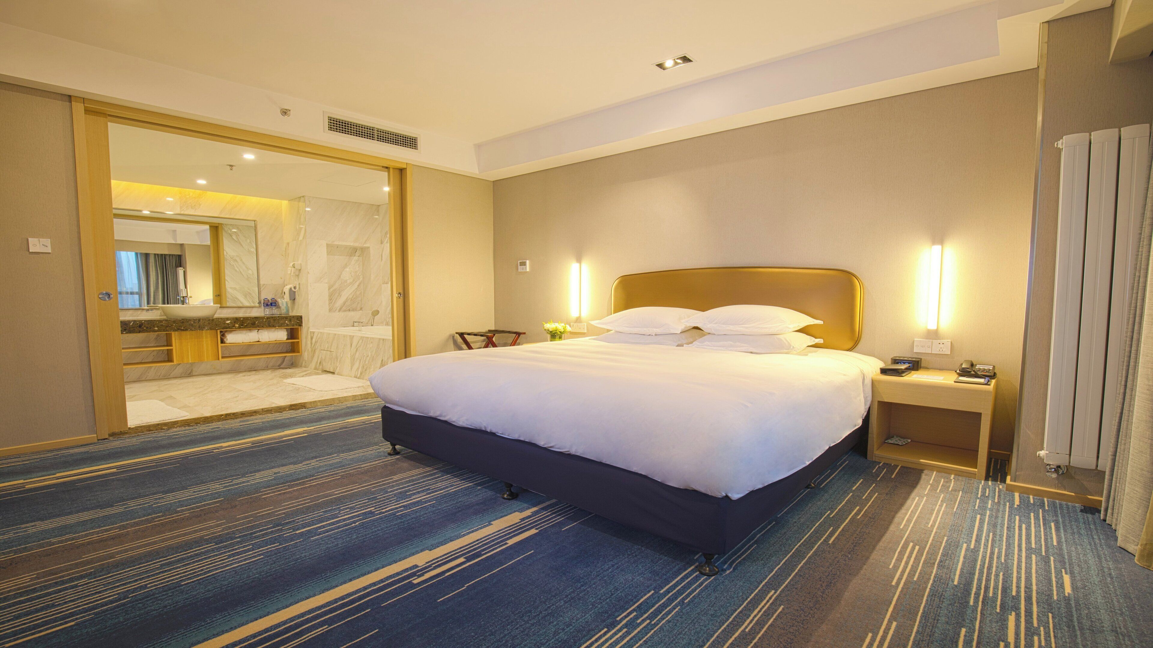A hotel room with a large bed, a bathroom with a mirror, and a nightstand with a lamp.